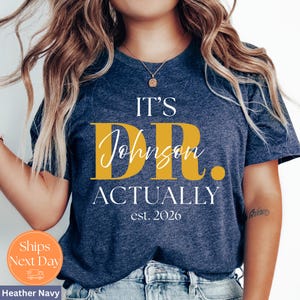 May include: Heather navy t-shirt with the text "IT'S DR. Johnson ACTUALLY est. 2026" in white and gold lettering. The shirt has a round neck and short sleeves. A small orange circle with the words "Ships Next Day" is visible.