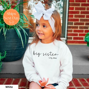 May include: White sweatshirt with the text "big sister" and the name "Lily Ann" in black script.