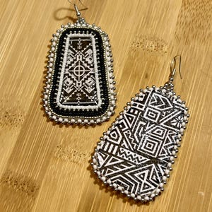May include: Two handmade earrings with intricate geometric patterns. The earrings feature black and white designs, with a beaded border in white and silver. The rectangular earring has a central design, while the other has a more abstract pattern.