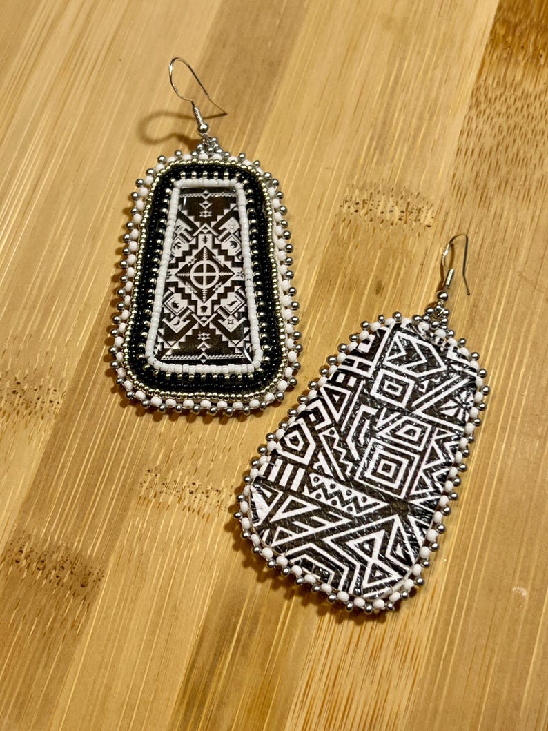 May include: Two handmade earrings with intricate geometric patterns. The earrings feature black and white designs, with a beaded border in white and silver. The rectangular earring has a central design, while the other has a more abstract pattern.