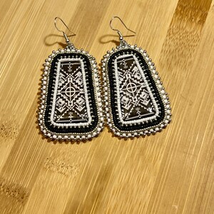May include: A pair of beaded earrings with a geometric design. The earrings feature a black and white pattern within a rectangular shape, framed by white and black beads. Silver-colored ear wires complete the design.
