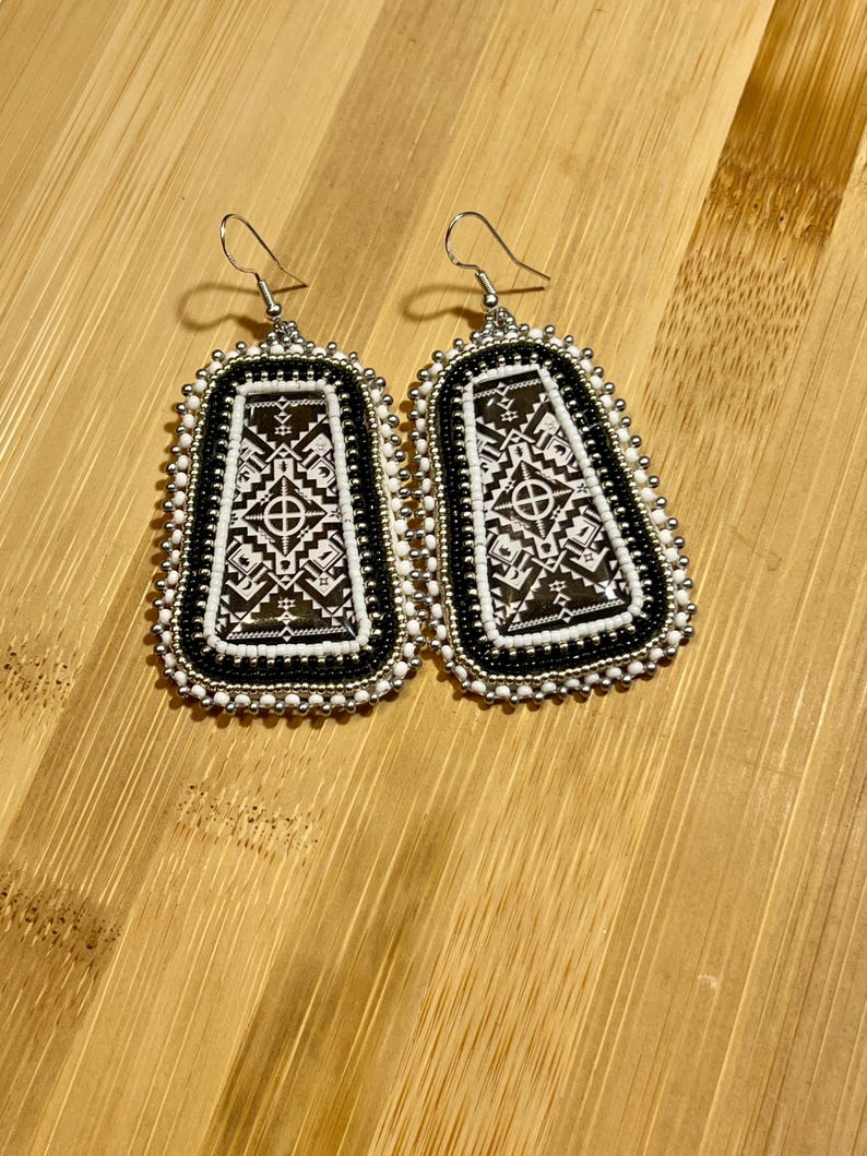 May include: A pair of beaded earrings with a geometric design. The earrings feature a black and white pattern within a rectangular shape, framed by white and black beads. Silver-colored ear wires complete the design.