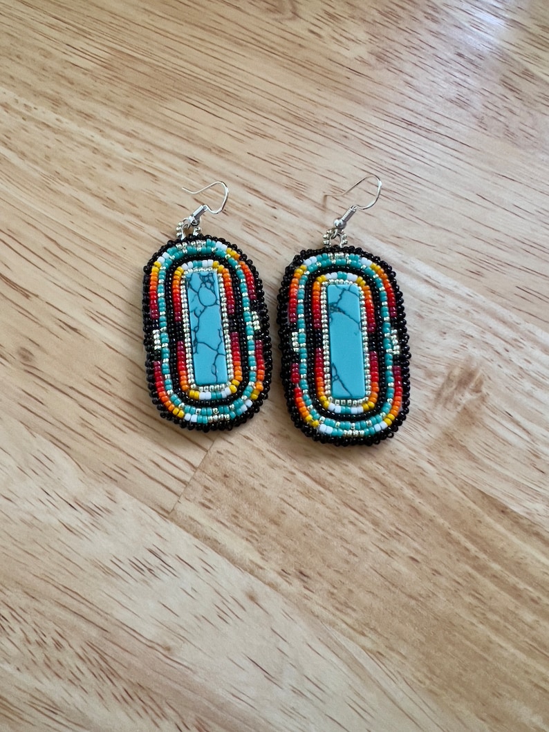 May include: A pair of turquoise and multi-colored beaded earrings with silver hooks. The earrings are oval shaped and feature a turquoise stone in the center.