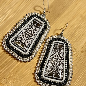 May include: A pair of beaded earrings with a geometric design. The earrings feature a black and white pattern with silver-colored accents. The earrings are rectangular with rounded edges and have a hook closure.