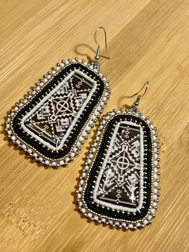 May include: A pair of beaded earrings with a geometric design. The earrings feature a black and white pattern with silver-colored accents. The earrings are rectangular with rounded edges and have a hook closure.
