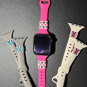 May include: A bright pink smartwatch band with a beaded geometric design in white, black, and pink. Two additional bands are shown: one gray with turquoise and white beading, and one cream-colored with pink and white beading. The bands are for a smartwatch.
