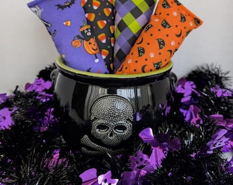 May include: A black plastic cauldron featuring a textured skull and crossbones design on the front, filled with four rectangular fabric pouches. The pouches display patterns of purple with cats and bats, black with candy corn, purple and green plaid, and orange with black cats and moons.