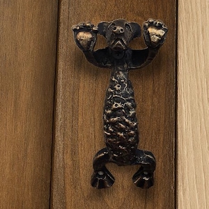 May include: A bronze cabinet pull in the form of a stylized animal. It's mounted on a dark brown wooden cabinet door. The animal figure has a textured body and is holding its arms up. The cabinet door is made of wood.