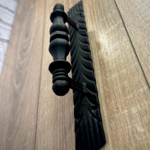 Może przedstawiać: Czarny metalowy klamka do drzwi z ozdobną, fakturowaną płytką. Klamka ma zaokrąglony, ozdobny wzór.