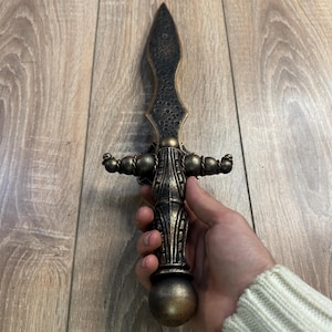 Op de afbeelding: Een decoratief dolk met een zwarte en gouden afwerking. De dolk heeft een gedetailleerd handvat met een ronde knop en een puntig blad.