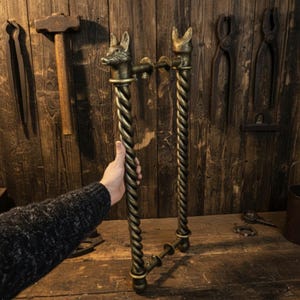 Puede incluir: Manijas de puerta de bronce ornamentadas con un diseño retorcido y remates de cabeza de zorro. Las manijas son sostenidas por una persona frente a un fondo de madera rústica con herramientas de herrero.