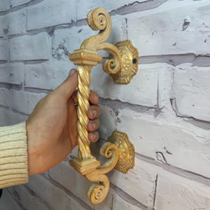 May include: A gold-painted, ornate door handle with a twisted design. The handle is mounted on a white brick wall.