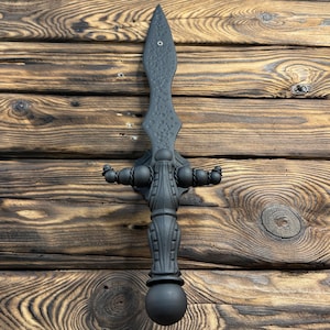 Puede incluir: Un gancho de pared de metal negro con forma de espada con una hoja texturizada y una bola redonda en la parte inferior. El gancho tiene tres puntas en el lateral para colgar objetos.