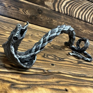 Könnte beinhalten: Ein silberner, drachenförmiger Türgriff aus Metall mit einem eingerollten Schwanz. Der Griff ist auf einer Holzoberfläche montiert.