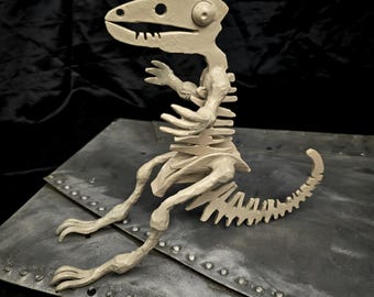 May include: A beige ceramic dinosaur skeleton sculpture with a detailed ribcage and tail. The sculpture is sitting on a metal surface with rivets.