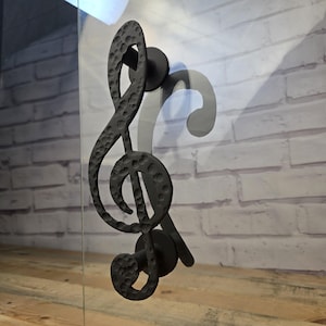 May include: A black metal door handle shaped like a treble clef. The handle has a textured surface and is mounted on a glass door. A white brick wall is visible in the background. The handle has a hammered appearance.