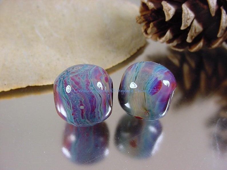 May include: Two round glass beads with a swirling pattern of blue, purple, and pink. The beads are iridescent and have a glossy finish.