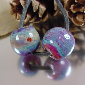 May include: Two colorful glass beads with swirls of blue, purple, and red, with white accents. The beads are strung on a black cord.