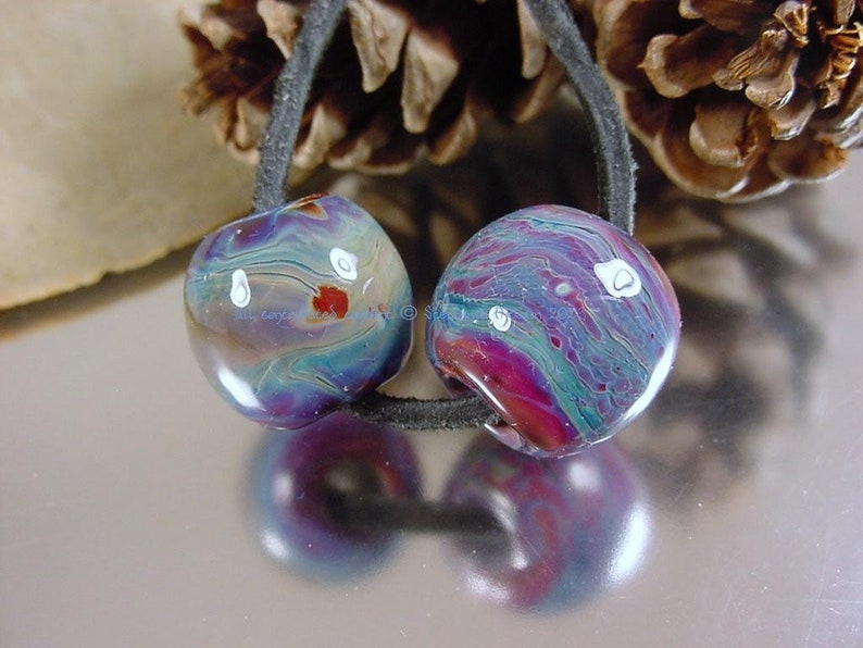 May include: Two colorful glass beads with swirls of blue, purple, and red, with white accents. The beads are strung on a black cord.