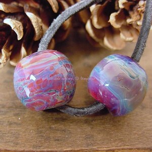 May include: Two colorful glass beads with a swirling pattern of pink, purple, and blue. The beads are strung on a black cord.