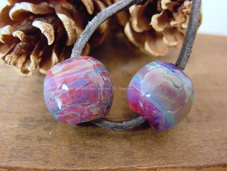 May include: Two colorful glass beads with a swirling pattern of pink, purple, and blue. The beads are strung on a black cord.
