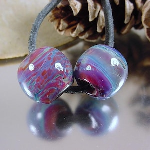May include: Two glass beads with a marbled pattern in shades of purple, blue, and red with white accents. The beads are strung on a black cord.