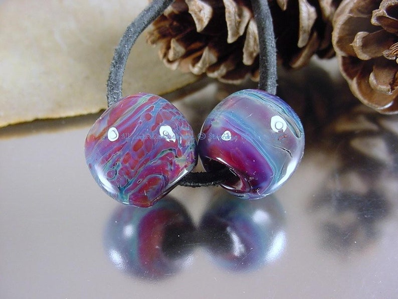 May include: Two glass beads with a marbled pattern in shades of purple, blue, and red with white accents. The beads are strung on a black cord.