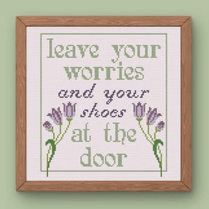 May include: A framed cross-stitch artwork with the text "leave your worries and your shoes at the door." The text is in green and purple, with floral accents. The frame is made of wood.