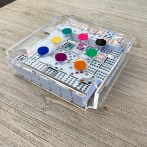 May include: A clear acrylic domino tray with a set of white dominoes inside. The dominoes are arranged in a pattern and have colorful dots on them. The tray has a raised edge to keep the dominoes from falling out.