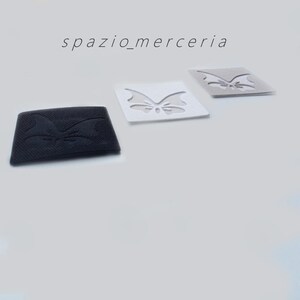 Può includere: Tre toppe in tessuto a forma di farfalla in nero, bianco e grigio. La toppa nera è rettangolare con un disegno a farfalla in rilievo. Le toppe bianca e grigia sono quadrate con un ritaglio a farfalla. Il testo "spazio_merceria" è in alto.