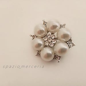 Może przedstawiać: Okrągła broszka w kolorze srebrnym z sześcioma dużymi sztucznymi perłami ułożonymi wokół grupy mniejszych, przezroczystych kryształków. Broszka ma kwiatowy wzór z misternymi detalami. Widoczny tekst "spazio_merceria". Białe tło.
