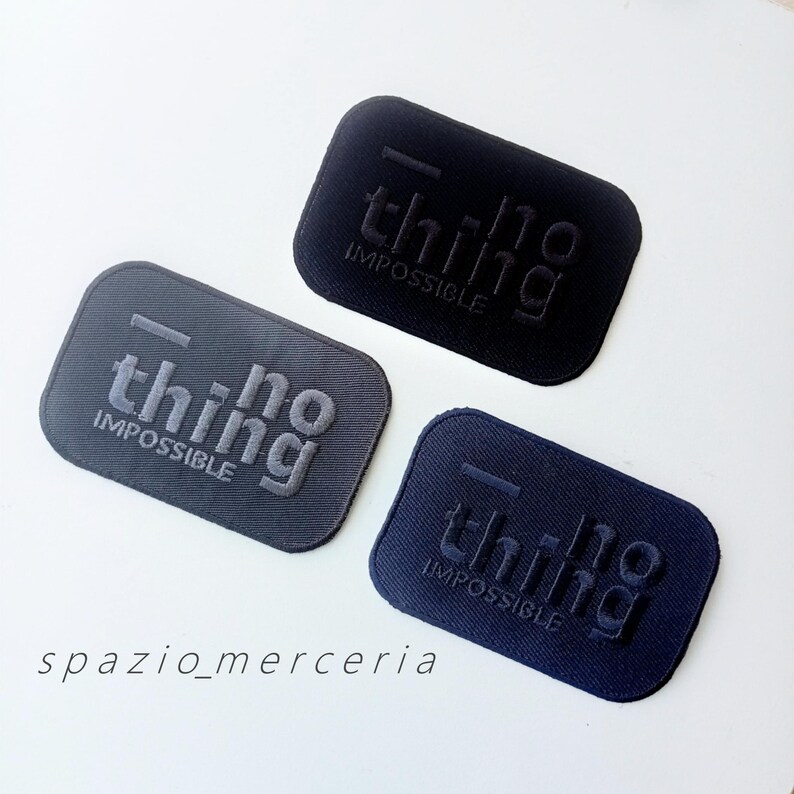 May include: Three rectangular fabric patches in black, gray, and navy blue. Each patch features the embossed words "thing impossible" in a stylized font. The patches are arranged on a white surface.