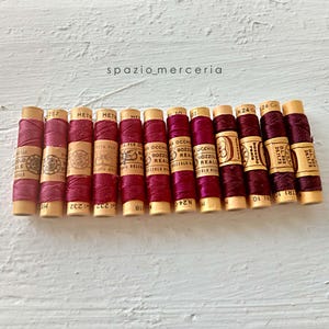 Può includere: Una fila di dodici rocchetti di legno vintage avvolti in filo color magenta intenso, ognuno con un'etichetta di carta color cuoio con il testo Bozzolo Reale e vari numeri. I rocchetti sono disposti orizzontalmente, mostrando una tonalità rosa scuro.