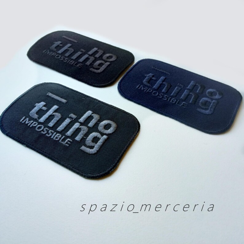 May include: Three rectangular patches with the words "no thing impossible" embroidered on them. The patches are in shades of black and navy blue. The text is in a contrasting color, and the patches are arranged on a white surface.