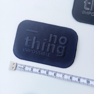 May include: Navy blue rectangular patch with the words "nothing impossible" embossed in a darker shade. A measuring tape is placed near the patch, showing centimeters. The patch has rounded corners and a textured surface.