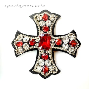 May include: Embroidered cross applique with red and clear rhinestones on a black felt base. The cross features a large, rectangular red stone at the center and is embellished with gold-colored thread. The text "spazio_merceria" is in the upper left corner.