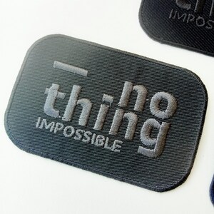 May include: A dark gray embroidered patch with the word "thing" above "IMPOSSIBLE" in gray thread. The patch has a rectangular shape with rounded corners. The text is in a stylized font.