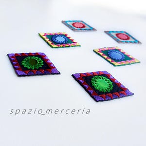 May include: Embroidered patches in various colors, including purple, red, green, and blue, arranged on a white surface. Each patch features a central design surrounded by a geometric border. The text "spazio_merceria" is visible at the bottom.
