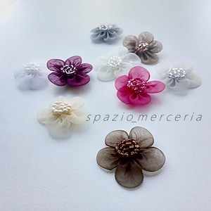 Op de afbeelding: Een verzameling kleine stoffen bloemversieringen in verschillende kleuren, waaronder wit, roze, paars en bruin. Elke bloem heeft een kralen- of parelachtig centrum. De tekst "spazio_merceria" is zichtbaar.