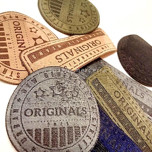May include: A selection of fabric labels in brown, green, grey, and blue. The labels display the word "ORIGINALS" in a distressed font, alongside other text and star designs. Some labels are oval, while others are rectangular.