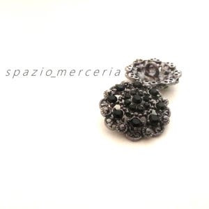 May include: Two ornate, antique-style buttons. The buttons are silver-toned with a floral design and are embellished with black gemstones. The text "spazio_merceria" is printed on the left side of the image.