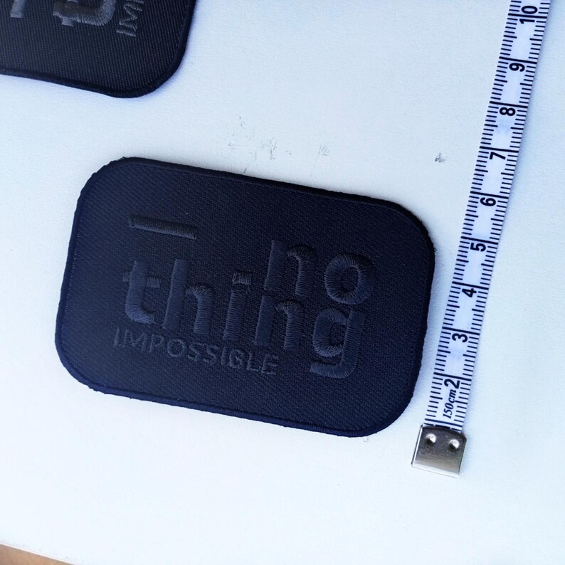 May include: Two black rectangular patches with the words "no thing impossible" embossed on them. A white and blue measuring tape is next to one of the patches, showing centimeters and inches.