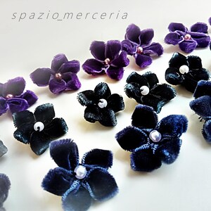 Può includere: Primo piano di abbellimenti floreali in velluto nei toni del viola, blu navy e nero, ciascuno con un centro di perla. I fiori sono disposti su una superficie bianca. Il testo "spazio_merceria" è visibile.