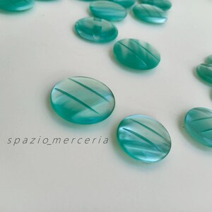 Könnte beinhalten: Nahaufnahme von mehreren ovalen, durchscheinenden türkisfarbenen Knöpfen mit diagonalen, geätzten Linien. Die Knöpfe sind auf einer weißen Oberfläche verstreut. Der Text "spazio_merceria" ist sichtbar. Ideal für Näh- und Bastelprojekte.