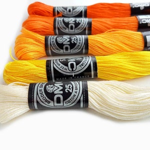 Può includere: Primo piano di diversi rocchetti di filo da ricamo DMC nei toni dell'arancione, giallo e crema. Ogni rocchetto ha un'etichetta in bianco e nero con il logo e il testo DMC. I fili sono disposti per mostrare i colori vivaci.