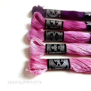 May include: A collection of DMC embroidery floss in shades of pink and purple. The floss is neatly wound on spools with black and silver labels. The image is well-lit, showcasing the vibrant colors and textures of the thread.