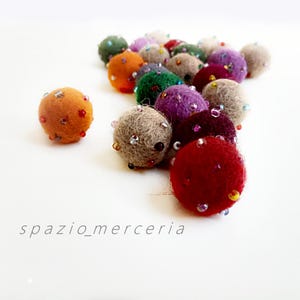Peut inclure: Un ensemble de boules en feutre multicolores, notamment orange, vert, violet et rouge, chacune ornée de petites perles scintillantes. Les boules sont disposées sur un fond blanc, avec le texte "spazio_merceria" en bas.