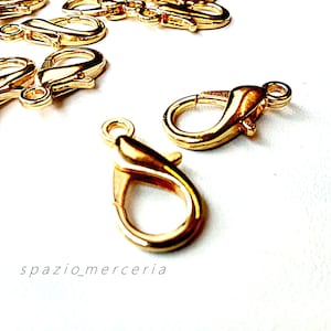 Può includere: Diversi moschettoni color oro sono sparsi su una superficie bianca. I moschettoni sono lucidi e hanno una finitura liscia. Il testo "spazio_merceria" è stampato in corsivo in basso nell'immagine. Questi moschettoni sono ideali per la creazione di gioielli.