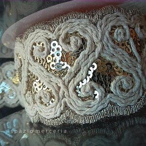 May include: A decorative trim ribbon featuring a beige corded rope pattern arranged in swirling loops. The design is accented with metallic gold sequins embedded within the mesh background, finished with a gold metallic border along the edges.