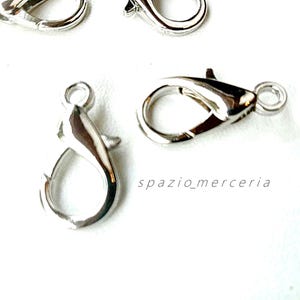 Può includere: Primo piano di diverse chiusure a moschettone color argento, comunemente utilizzate nella creazione di gioielli e nell'artigianato. Le chiusure sono disposte su una superficie bianca. Il testo "spazio_merceria" è visibile.